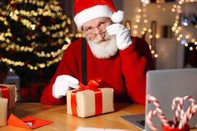 Photo of Santa Claus tying bow on gift box at his workplace in room decorated for Christmas