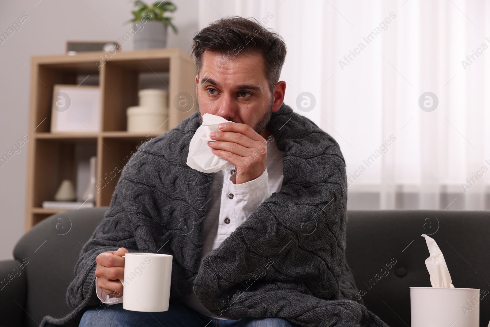 Photo of Sick man wrapped in blanket with cup of drink and tissue blowing nose on sofa at home. Cold symptoms