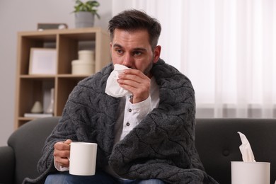 Sick man wrapped in blanket with cup of drink and tissue blowing nose on sofa at home. Cold symptoms