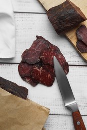 Photo of Pieces of delicious beef jerky and knife on white wooden table, flat lay