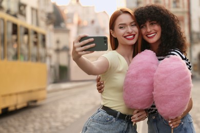 Photo of Happy friends with pink cotton candies taking selfie on city street. Space for text