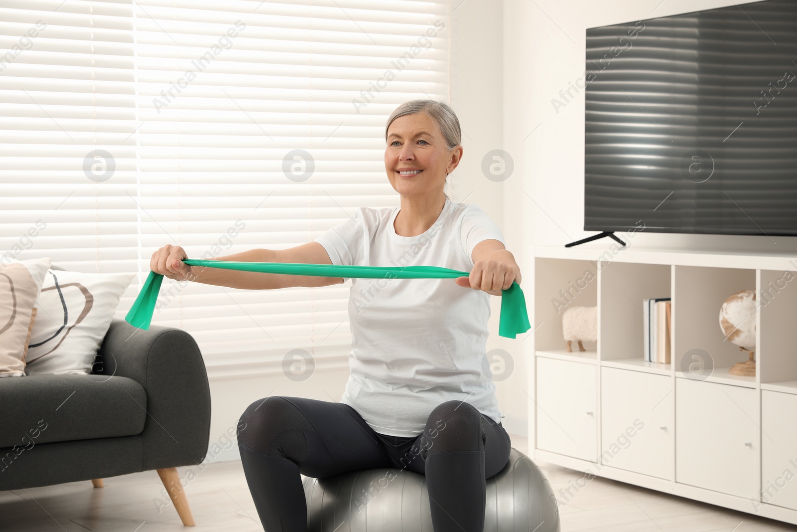 Photo of Senior woman doing exercise with elastic resistance band on fitness ball at home