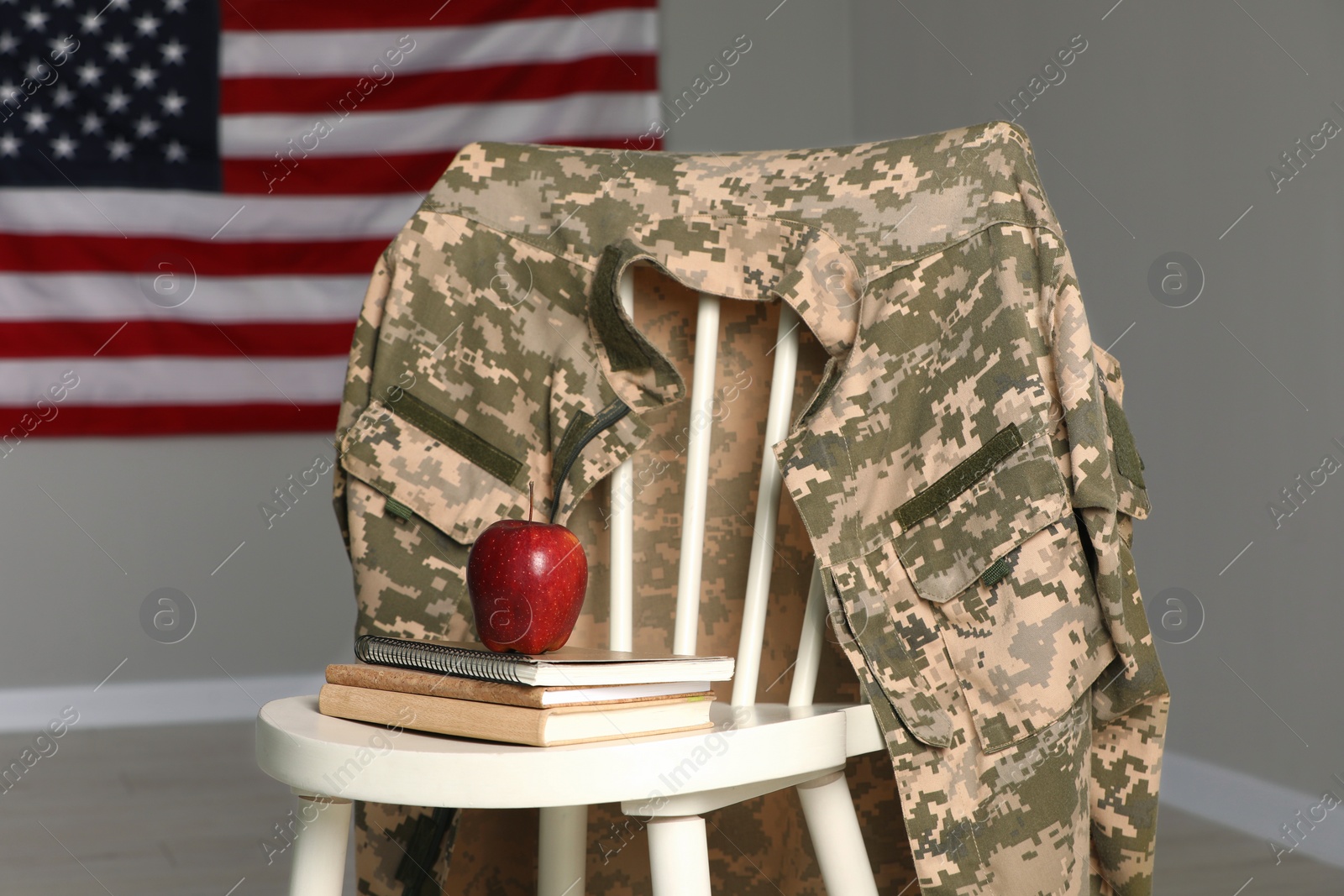 Photo of Chair with soldier uniform, notebooks and apple near flag of United States indoors. Military education