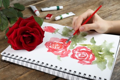 Photo of Woman painting roses in sketchbook at wooden table, closeup