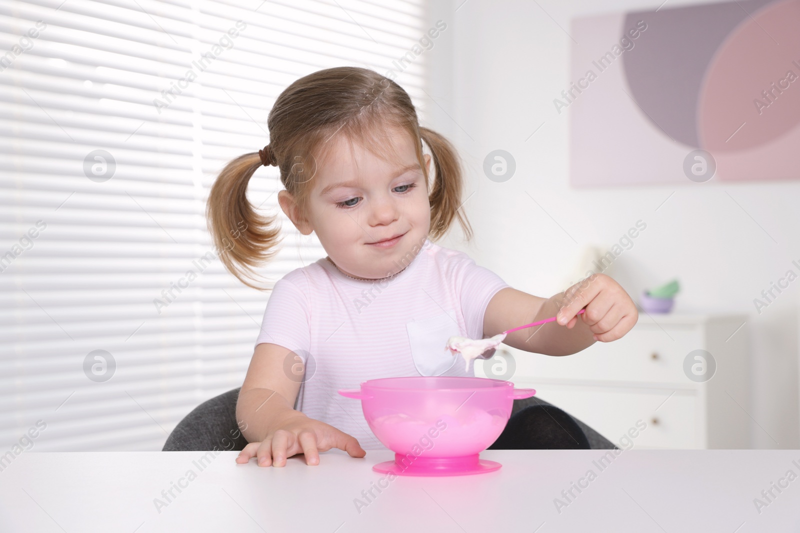 Photo of Cute little child eating tasty yogurt from plastic bowl with spoon at white table indoors