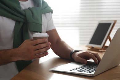 Freelancer with cup of coffee working on laptop at table indoors, closeup