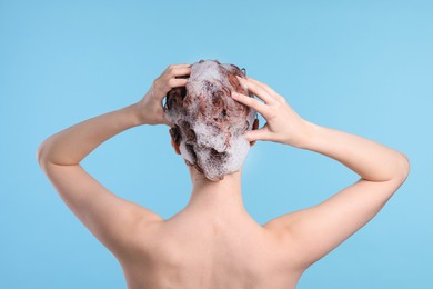 Young woman washing her hair with shampoo on light blue background, back view