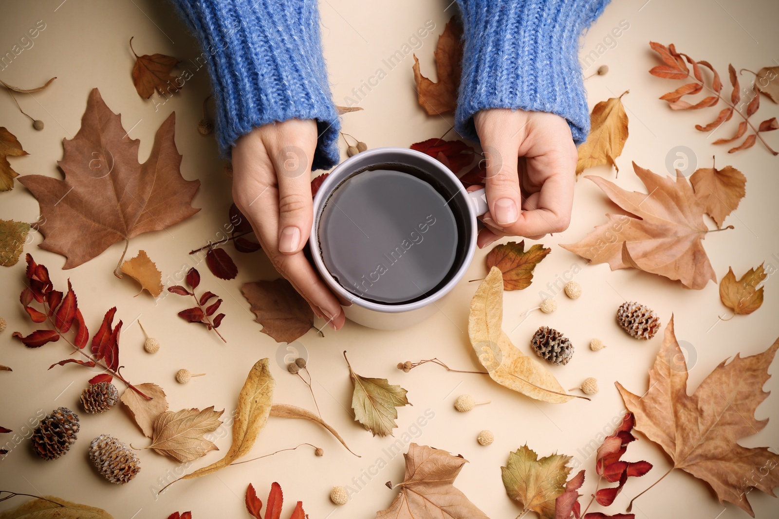Photo of Woman with cup of hot drink surrounded by autumn leaves on beige background, flat lay. Cozy atmosphere