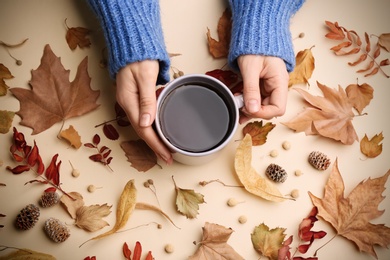 Woman with cup of hot drink surrounded by autumn leaves on beige background, flat lay. Cozy atmosphere