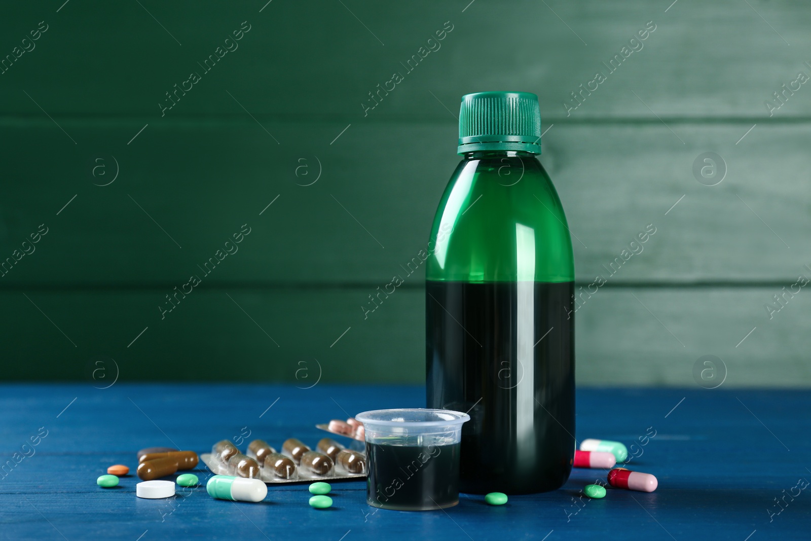 Photo of Bottle of cough syrup, measuring cup and pills on blue wooden table