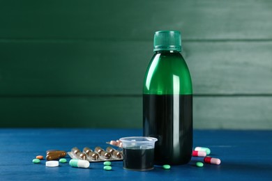 Bottle of cough syrup, measuring cup and pills on blue wooden table