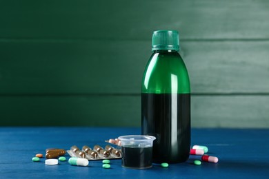 Photo of Bottle of cough syrup, measuring cup and pills on blue wooden table