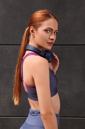 Beautiful woman in stylish gym clothes with headphones posing near dark grey wall on street