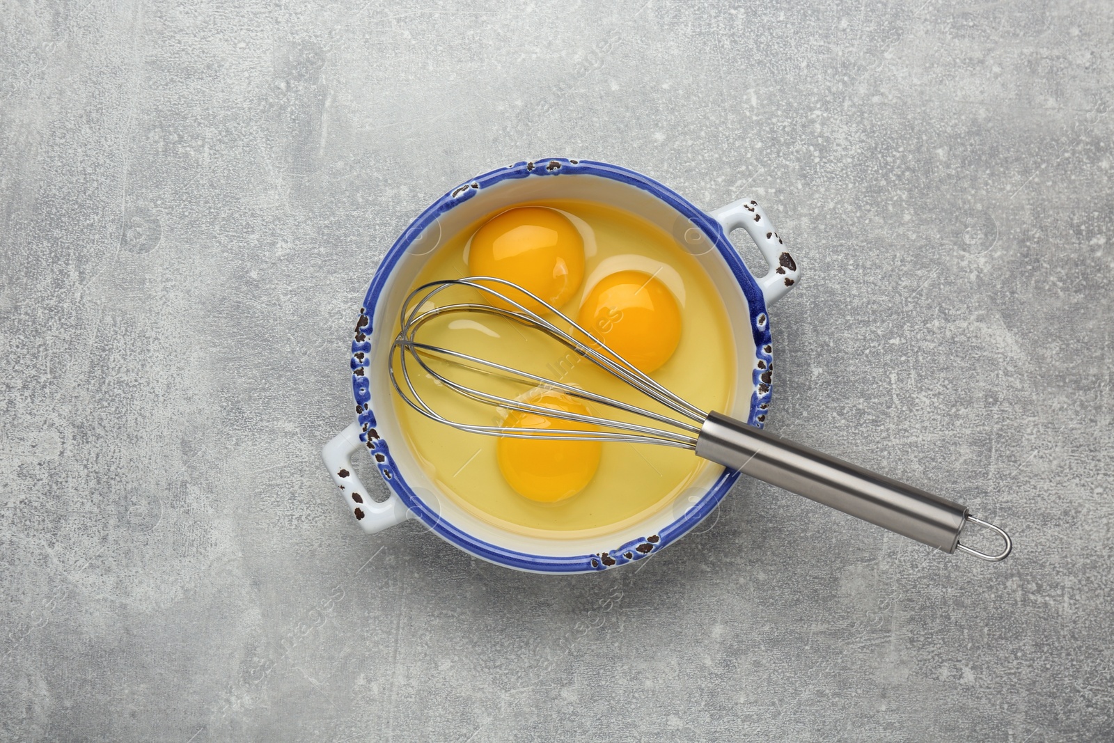 Photo of Whisk and eggs in pot on grey table, top view