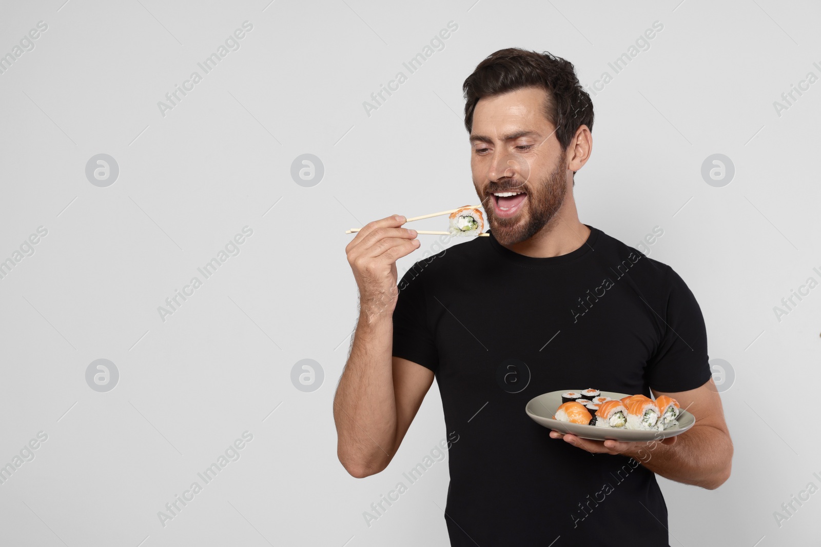 Photo of Happy man eating tasty sushi roll with chopsticks on light grey background. Space for text