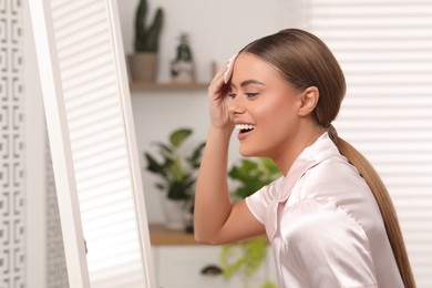 Beautiful woman removing makeup with cotton pad near mirror indoors