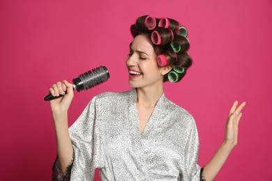 Beautiful young woman in silk bathrobe with hair curlers singing into hairbrush on pink background