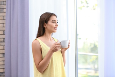 Young woman with cup of drink near window with open curtains at home