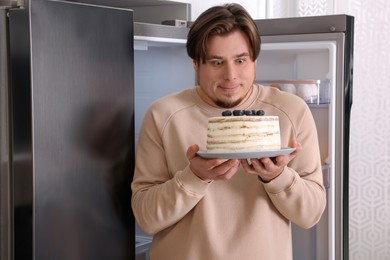 Photo of Happy overweight man with cake near open refrigerator in kitchen