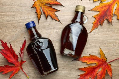 Photo of Glass bottles of tasty maple syrup and dry leaves on wooden table, flat lay