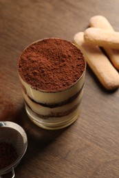 Photo of Delicious tiramisu in glass, biscuits and sieve with cocoa powder on wooden table