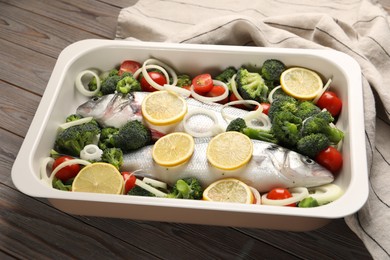 Raw fish with vegetables and lemon in baking dish on wooden table