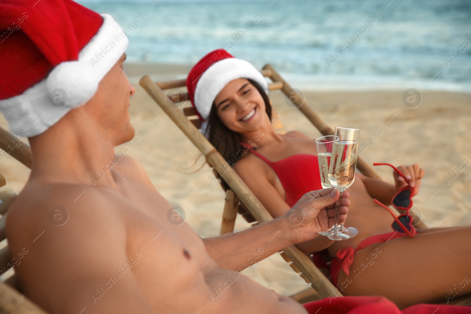 Photo of Happy couple wearing Santa hats and drinking champagne together on beach, focus on glasses. Christmas vacation