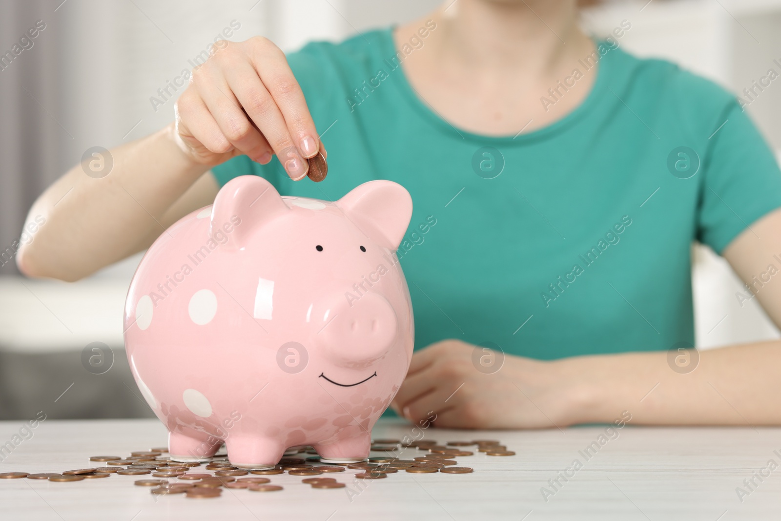 Photo of Financial savings. Woman putting coin into piggy bank at white wooden table indoors, closeup