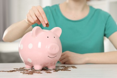 Financial savings. Woman putting coin into piggy bank at white wooden table indoors, closeup