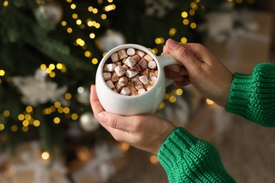 Photo of Woman holding cup of delicious Christmas cocoa with marshmallows near fir tree, above view