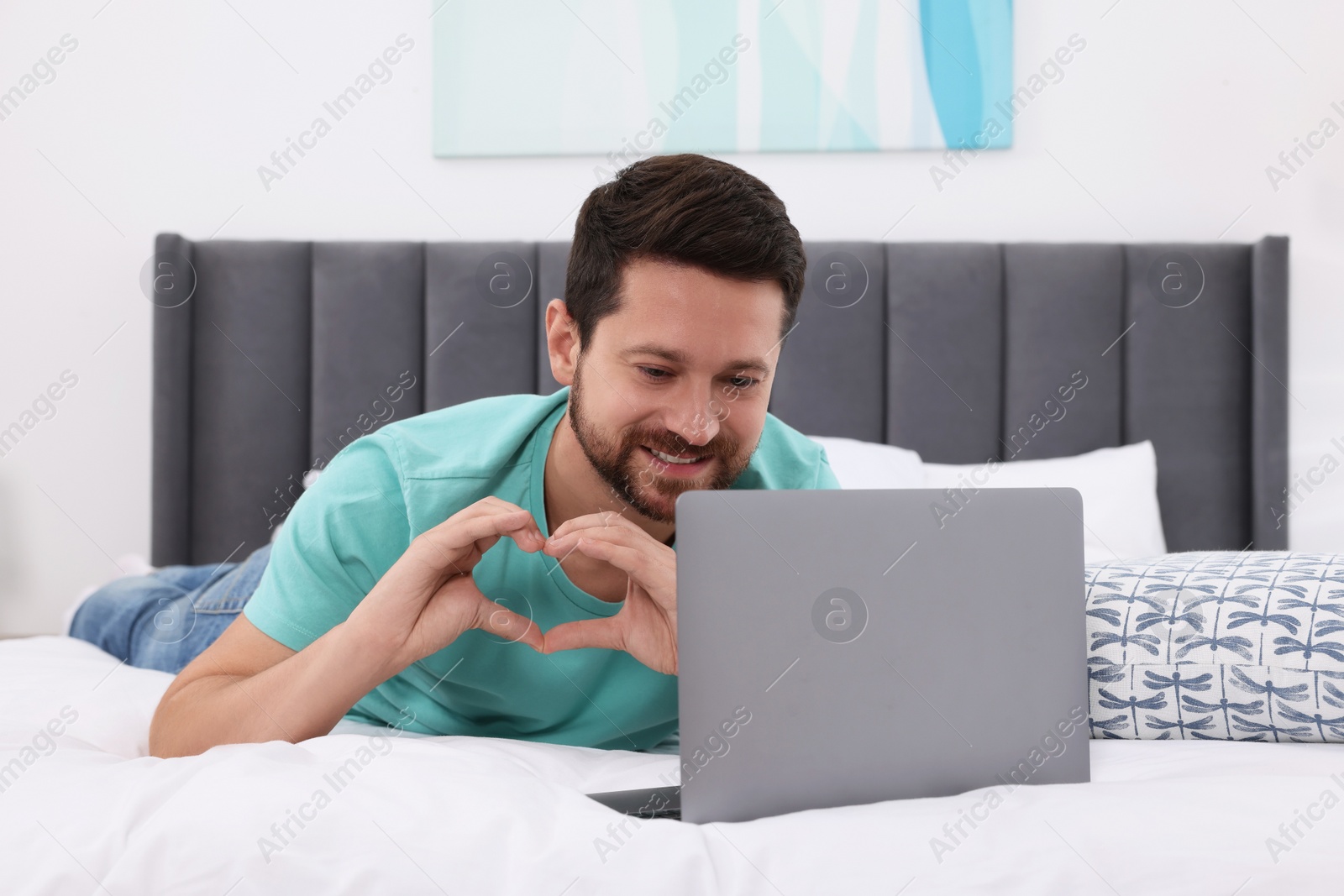 Photo of Happy man making heart with hands during video chat via laptop in bedroom. Long-distance relationship