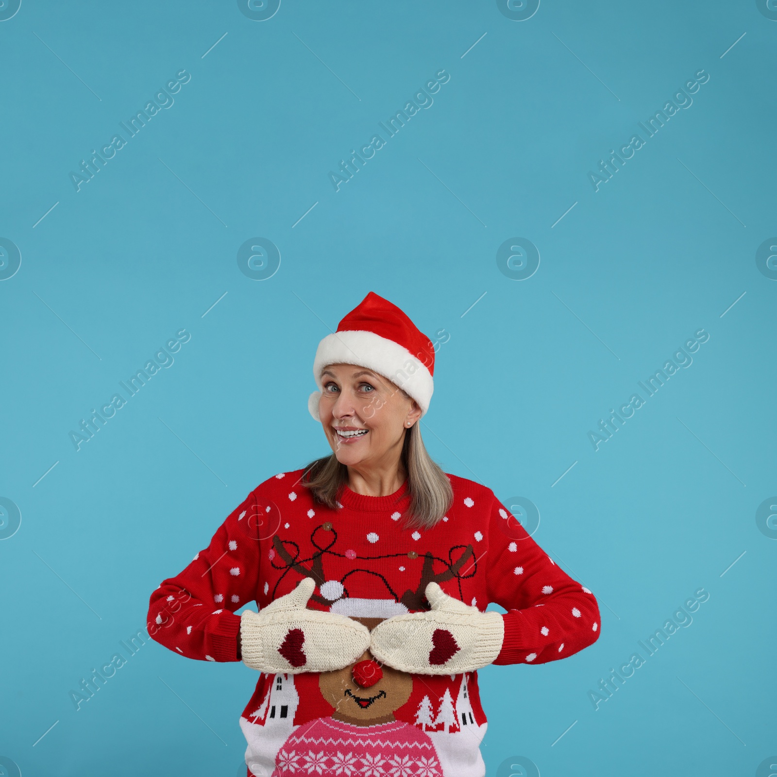 Photo of Happy senior woman in Christmas sweater, Santa hat and knitted mittens on light blue background