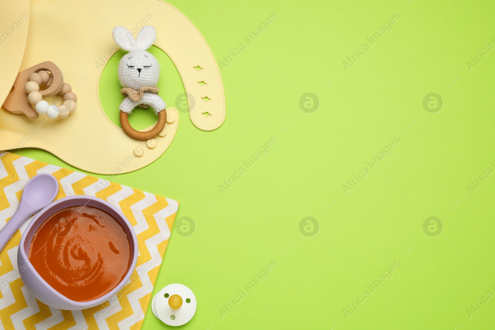 Photo of Silicone baby bib and plastic dishware with healthy food on light green background, flat lay. Space for text