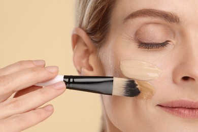 Photo of Woman applying foundation on face with brush against beige background, closeup