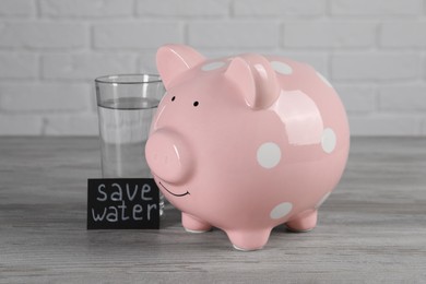 Photo of Card with phrase Save Water. Piggy bank and glass of drink on wooden table