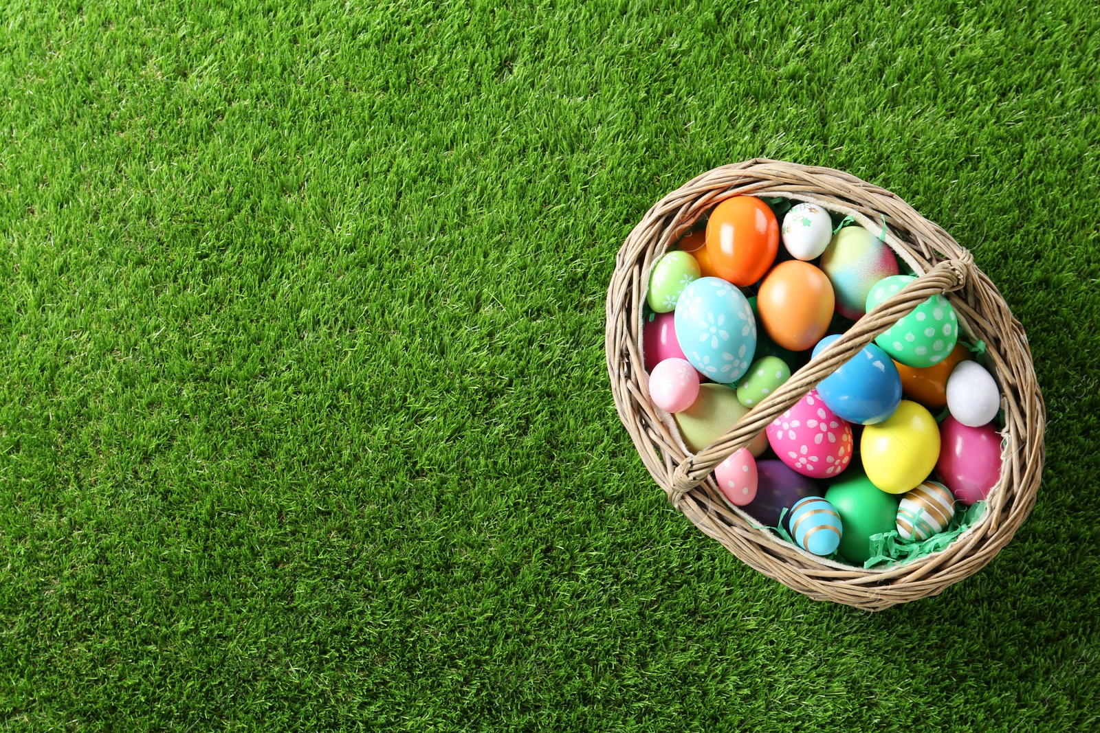 Photo of Wicker basket with Easter eggs on green grass, top view. Space for text