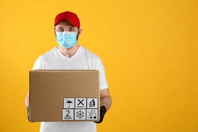 Courier in mask holding cardboard box with different packaging symbols on yellow background, space for text. Parcel delivery