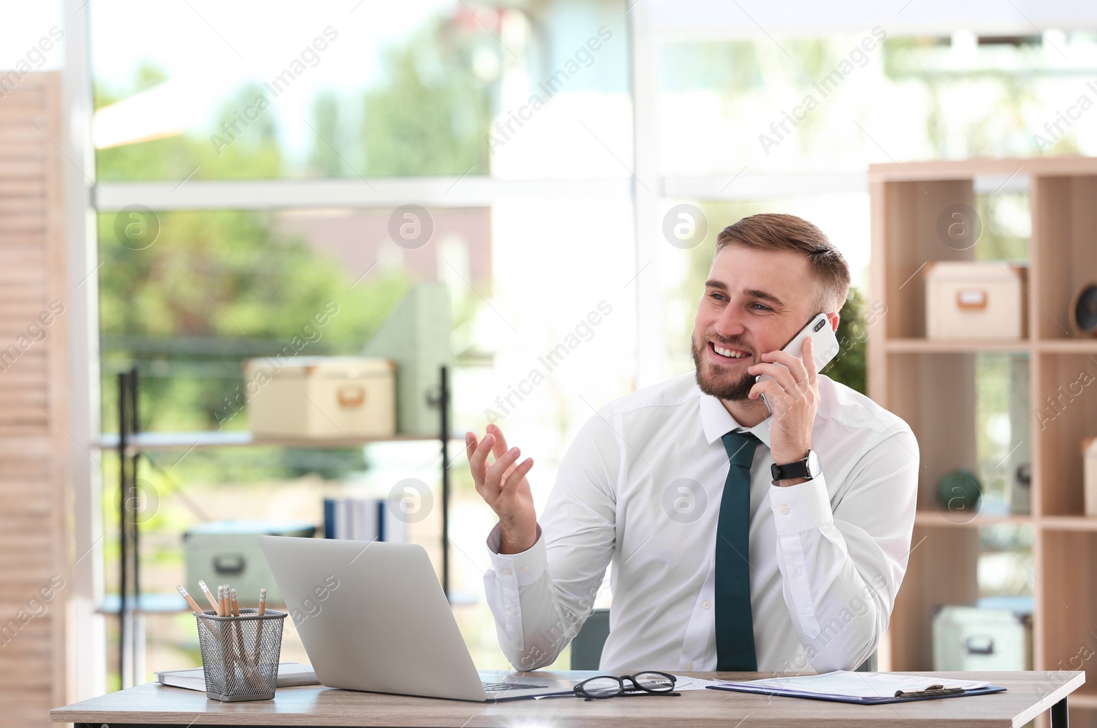 Photo of Young businessman talking on phone while using laptop at table in office