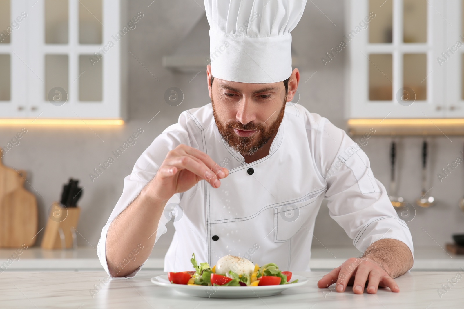 Photo of Professional chef salting delicious salad at marble table in kitchen