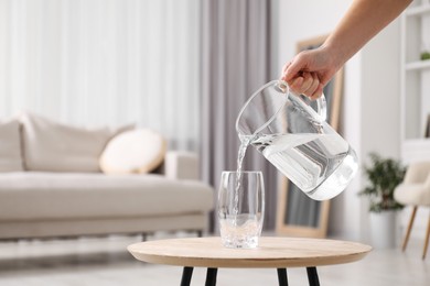 Woman pouring fresh water from jug into glass at wooden table indoors, closeup. Space for text