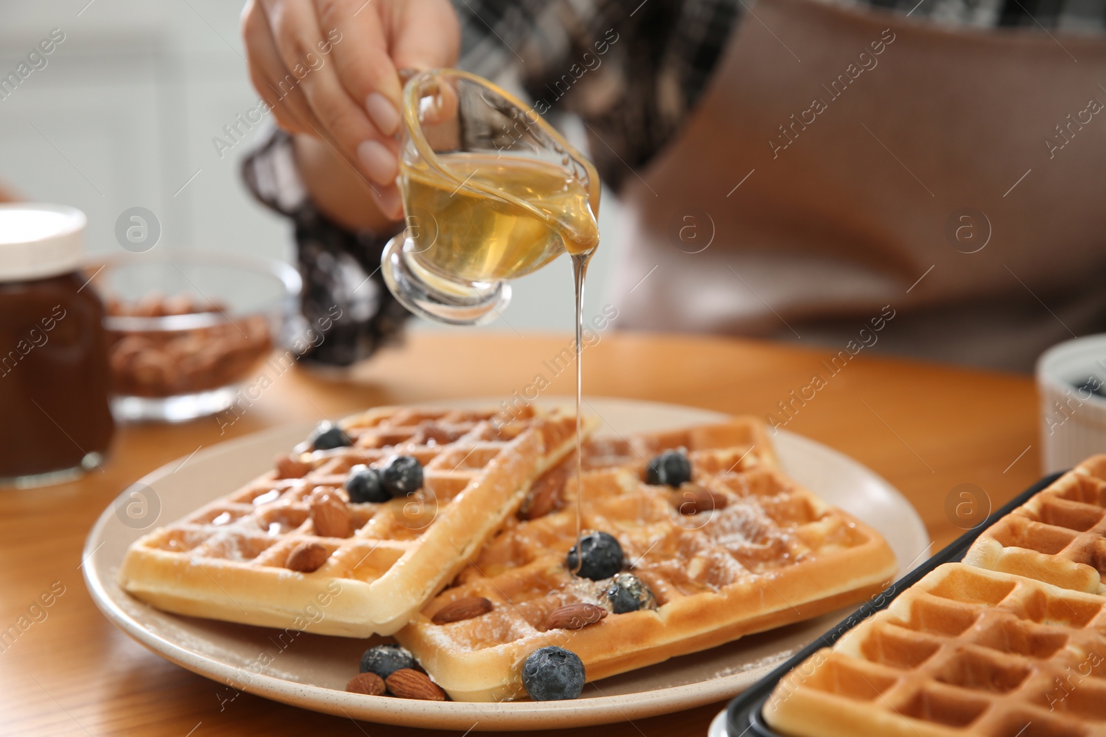 Photo of Woman pouring honey onto delicious Belgian waffles  at wooden table in kitchen, closeup