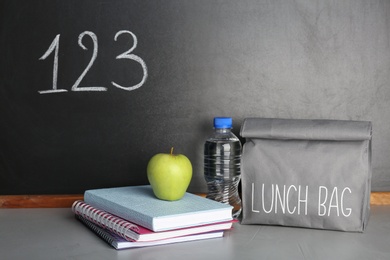 Photo of Healthy food for school child in lunch bag and stationery on table near blackboard with chalk written numbers