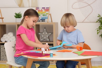 Photo of Girl using glue stick and boy cutting paper at desk in room. Home workplace