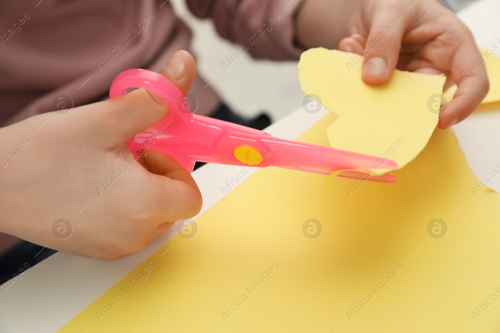 Photo of Child cutting out paper heart with plastic scissors at table, closeup