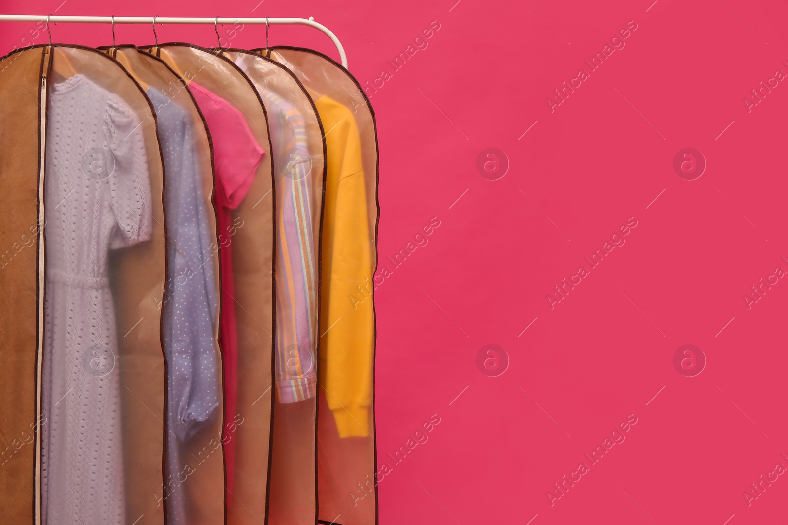 Photo of Garment bags with clothes on rack against pink background. Space for text