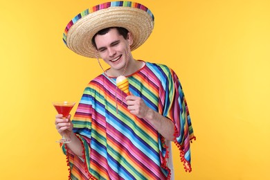 Young man in Mexican sombrero hat and poncho with cocktail and maracas on yellow background