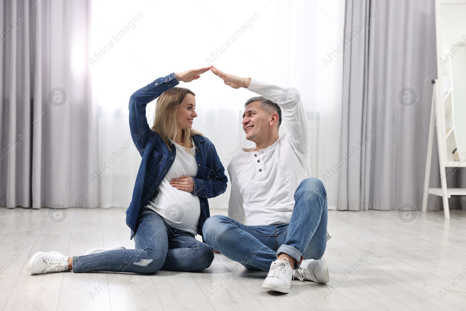 Photo of Young family housing concept. Pregnant woman with her husband forming roof with their hands while sitting on floor at home