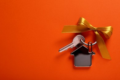 Key with trinket in shape of house and yellow bow on orange background, top view. Space for text. Housewarming party
