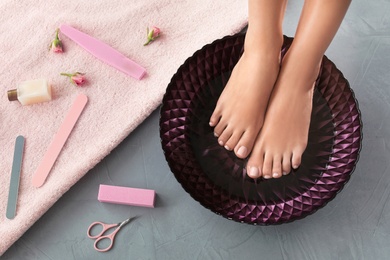 Photo of Woman soaking her feet in dish with water on grey floor, top view. Spa treatment