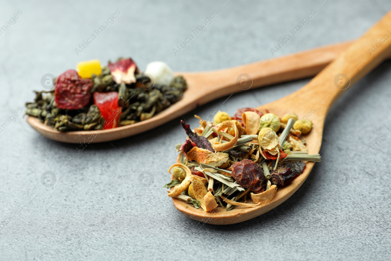 Photo of Different kinds of dry herbal tea in wooden spoons on light grey table, closeup
